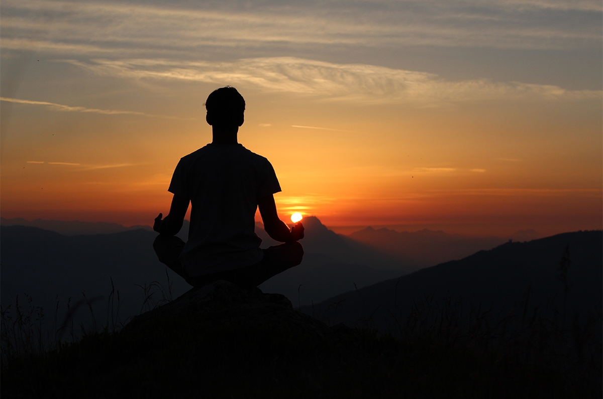 photo of person from behind sitting in lotus pose in front of the setting sun on a hilltop